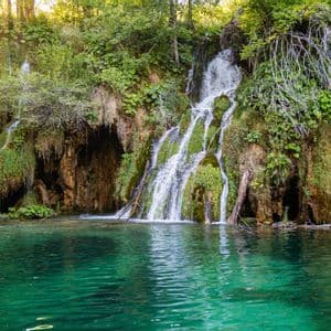 Wasserfälle stürzen einen moosbewachsenen Felsen hinab in einen türkisfarbenen See in einem sonnigen Wald.