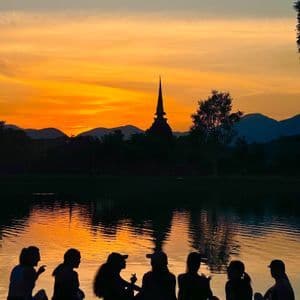 Eine WeRoad-Gruppe sitzt als Silhouette an einem See und beobachtet einen orangefarbenen Sonnenuntergang hinter einem fernen Tempel und Bergen.