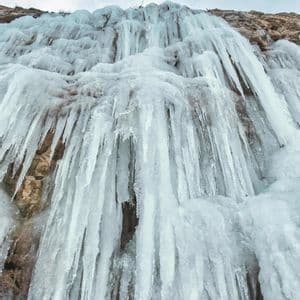 Eine Tiefenansicht eines gefrorenen Wasserfalls mit zahlreichen Eiszapfen, die an einer felsigen Klippenwand unter einem bewölkten Himmel hängen.