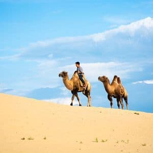 Eine Person reitet ein Kamel eine Sanddüne entlang, ein zweites Kamel begleitet sie, unter einem teilweise bewölkten blauen Himmel.