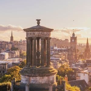 Un monumento classico con colonne su una collina erbosa che domina lo skyline di una città storica con guglie all'ora d'oro.