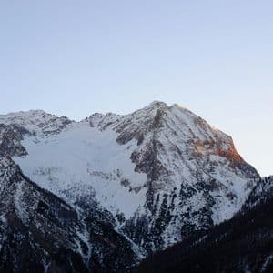 Un sommet enneigé est illuminé par la lumière orange du lever du soleil sous un ciel clair, avec des pentes boisées sombres en contrebas.