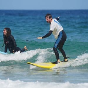 Un homme en voyage de groupe WeRoad fait de l'équilibre sur une planche de surf jaune tandis qu'une femme sur une autre planche sourit dans l'océan.
