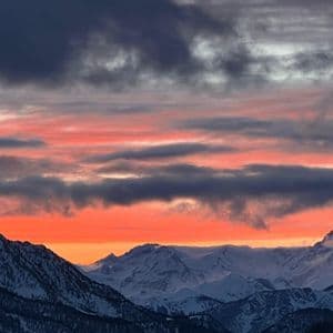 Ein dramatischer orange-roter Sonnenuntergang mit dunklen Wolken entfaltet sich über einer schneebedeckten Bergkette.