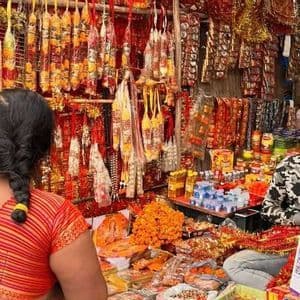 Una donna in sari osserva una bancarella in un mercato all'aperto, piena di ghirlande colorate e articoli religiosi venduti da un uomo.