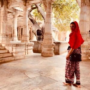 Una donna a piedi nudi con un foulard rosso si guarda indietro mentre si trova in un antico tempio con colonne di pietra intagliate e ornate.