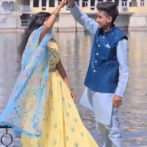 Un uomo e una donna in abiti tradizionali indiani ballano insieme, tenendosi per mano vicino a uno specchio d'acqua con un edificio sullo sfondo.