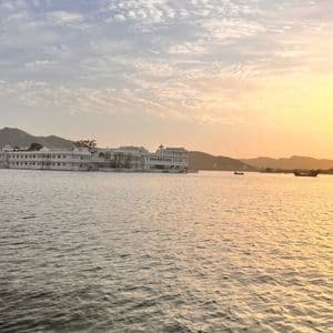 Un grande edificio bianco affacciato su un lago al tramonto, con colline lontane sotto un cielo parzialmente nuvoloso.