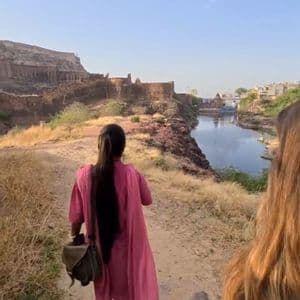 Due donne in un viaggio di gruppo WeRoad camminano su un sentiero sterrato lungo un fiume, con antiche fortificazioni in pietra sullo sfondo.