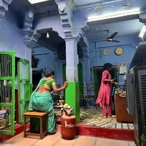 Due donne in una casa tradizionale con pareti e pilastri blu, una cucina seduta su uno sgabello vicino a una bombola del gas.