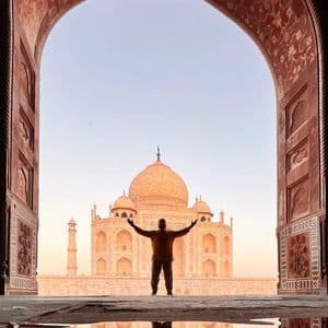 Una persona in silhouette a braccia alzate si erge davanti al Taj Mahal, visto attraverso un arco ornato con un riflesso in primo piano.