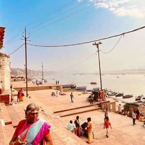 Una donna anziana in un sari colorato sale dei gradini di pietra che si affacciano su un ampio fiume con barche e persone sulla riva.