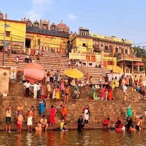 Una folla di persone vestite con abiti colorati si raduna su gradini di pietra accanto a un fiume, con alcuni che fanno il bagno nell'acqua.