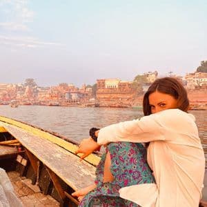 Una donna con pantaloni a motivi colorati siede in una barca di legno su un fiume, con uno skyline cittadino sullo sfondo.