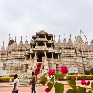 Un tempio in pietra ornato con una scalinata rivestita di tappeto rosso, con persone nel cortile e fiori rosa in primo piano.