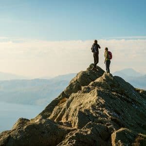 Dos excursionistas de un viaje en grupo de WeRoad están de pie en la cima rocosa de una montaña, contemplando un vasto paisaje de montañas y un lago.