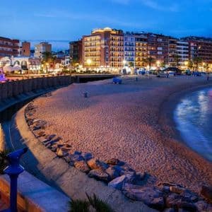 Una vista panoramica di una città di mare al tramonto, con edifici illuminati che costeggiano un'ampia spiaggia sabbiosa e acque calme dell'oceano.