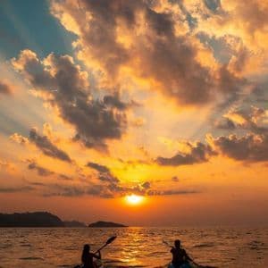 Due persone in kayak sul mare al tramonto, con i raggi del sole che filtrano tra nuvole arancioni e blu.