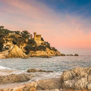Un castello si erge su una scogliera rocciosa e alberata che si affaccia su una spiaggia sabbiosa e sull'oceano durante un tramonto colorato con la luna nel cielo.