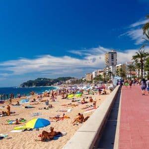 Una spiaggia affollata in una giornata di sole, con persone che prendono il sole sulla sabbia e passeggiano lungo un lungomare rosso costeggiato da palme.
