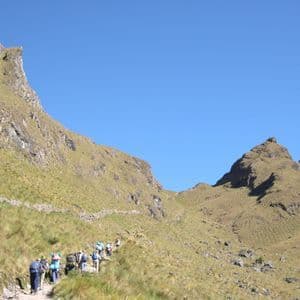 A WeRoad group trip with backpacks hiking in single file along a narrow trail on a grassy mountainside under a clear blue sky.