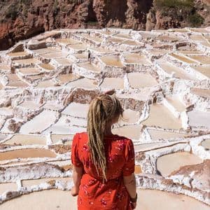 A woman with blonde dreadlocks in a red top, seen from behind, looking out over terraced salt evaporation ponds.