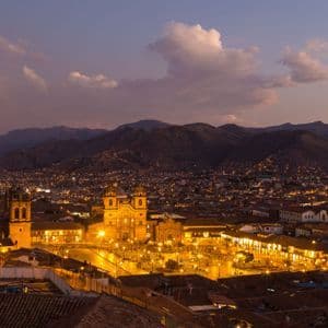 An illuminated city with a central plaza and cathedral at dusk, nestled in a valley surrounded by mountains.
