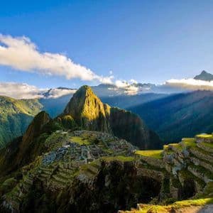 Ancient stone ruins and agricultural terraces sit on a verdant mountainside, illuminated by golden sunlight under a blue sky with clouds.