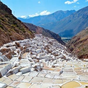 Terraced salt evaporation ponds fill a steep valley, with large mountains in the background under a partly cloudy sky.