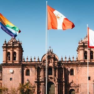 A rainbow flag, the flag of Peru, and the flag of Cusco waving on flagpoles in front of a large stone cathedral.