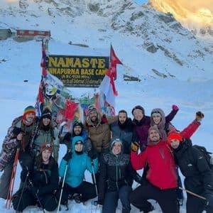 Un viaje en grupo de WeRoad posa para una foto en la nieve frente al cartel del Campo Base Annapurna, con un pico de montaña al fondo.