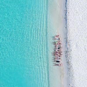 An aerial view of a WeRoad group trip lying in a line on a white sand beach beside clear turquoise water.