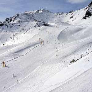 Una vista ampia di una pista da sci affollata con molte persone che sciano, circondata da montagne innevate e impianti di risalita.