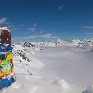 Un snowboard coloré se dresse dans la neige sur une montagne, surplombant une mer de nuages et des sommets enneigés sous un ciel bleu.