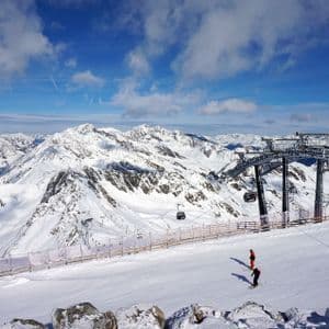 A wide view of a ski resort with two skiers on a slope, a gondola lift, and snow-capped mountains under a blue sky.