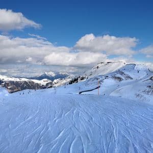 Una ladera de montaña cubierta de nieve con pistas de esquí en primer plano y esquiadores distantes bajo un cielo azul con nubes blancas.