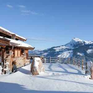 A wooden mountain cabin with a stone foundation is covered in snow, overlooking a snowy mountain range under a clear sky.
