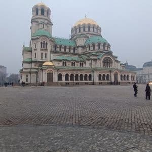 Una gran catedral con cúpulas doradas y verdes se alza frente a una amplia plaza empedrada en un día neblinoso.