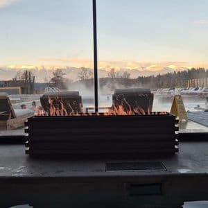 Una moderna hoguera arde junto a una humeante piscina termal exterior con bañistas, con un telón de fondo de montañas nevadas al anochecer.