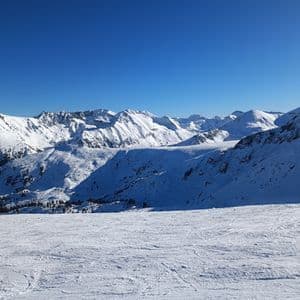 Una vista panorámica de una cordillera nevada bajo un cielo azul brillante, con una pista de esquí en primer plano.