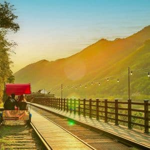 Una coppia percorre una bici su rotaia con una tettoia rossa lungo un binario attraverso una valle di montagna durante un tramonto dorato.