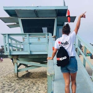 A WeRoad group leader seen from behind stands on a ramp next to a lifeguard tower on a sandy beach, pointing upwards.