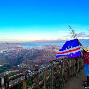 Eine Person mit Strohhut hält die Flagge Kap Verdes an einem Aussichtspunkt mit Blick auf eine Küstenstadt und Berge.