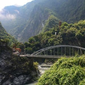 Un ponte ad arco in acciaio attraversa una gola fluviale rocciosa, circondato da montagne verdi e lussureggianti, con una pagoda visibile in lontananza.