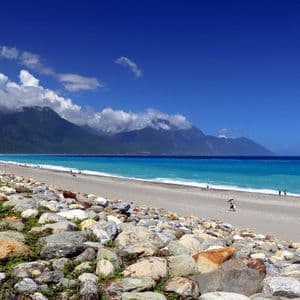 Una costa rocciosa conduce a una spiaggia sabbiosa lungo un mare turchese, con montagne verdi sotto un cielo azzurro parzialmente nuvoloso.
