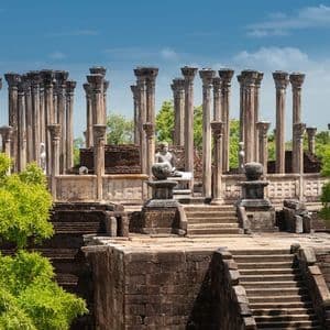 The ruins of an ancient temple with stone pillars and statues are framed by lush green trees under a blue sky.