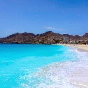 Türkisfarbene Meereswellen spülen an einen weißen Sandstrand vor einer Küstenstadt, umrahmt von kargen Bergen unter strahlend blauem Himmel.