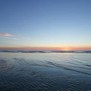 Die Sonne sinkt am Horizont über dem Ozean und wirft einen Schein auf den gewellten Sand eines feuchten Strandes bei Ebbe.