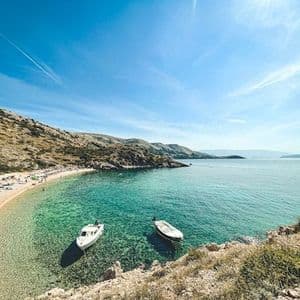 Eine Draufsicht auf eine sandige Bucht mit klarem, türkisfarbenem Wasser, zwei geankerten Booten und Personen am Strand.