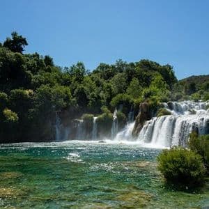 Ein mehrstufiger Wasserfall stürzt in einen klaren türkisfarbenen Fluss, umgeben von üppigem grünen Wald unter einem strahlend blauen Himmel.